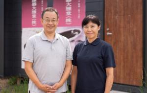 仮設工房の前でほほ笑む若島社長と妻の土百江さんの写真